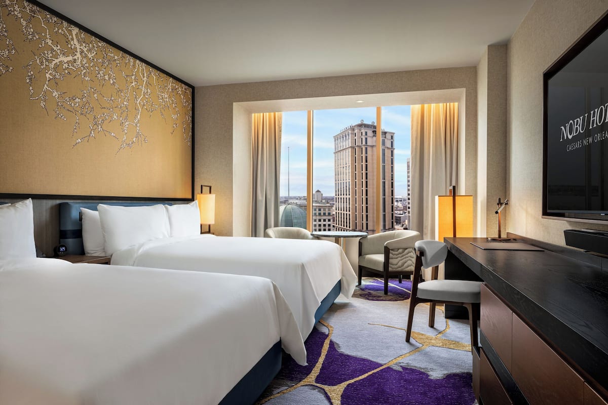 A hotel room with two beds, a desk, a chair, a TV, and a large window with a city view.