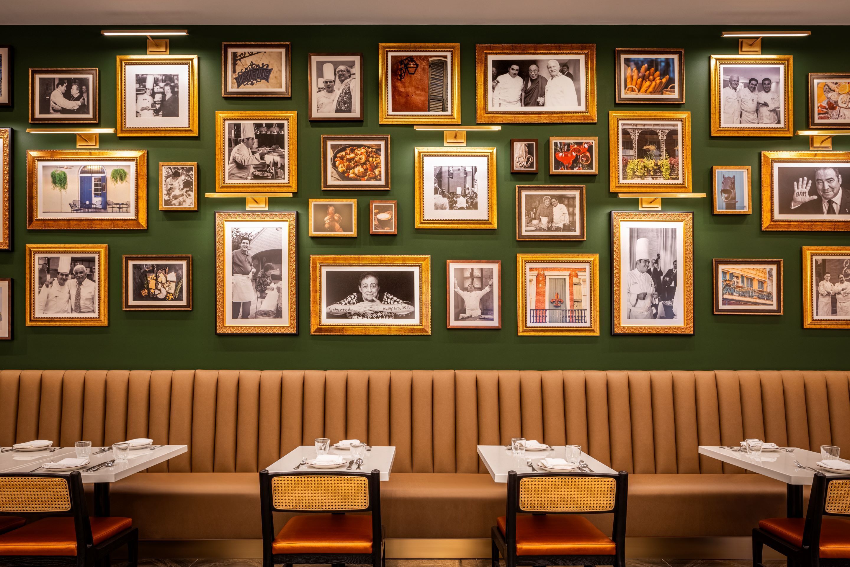 A restaurant with a green wall adorned with framed photographs, a brown booth seating area, and tables set with white tableware.