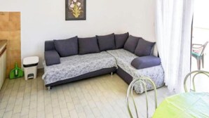 Una habitación con un sofá seccional gris, un cubo de basura blanco pequeño, una botella verde y una silla metálica.