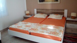 Una cama doble con una colcha naranja y sábanas estampadas con motivos florales, dos mesitas de noche con lámparas, un cuadro de una vista marina en la pared y una ventana con cortinas.
