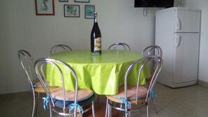 Un comedor con una mesa redonda cubierta con un mantel verde, rodeada de sillas de metal adornadas con cintas azules. Hay una botella de cerveza Zwickel sobre la mesa. En la pared hay cuatro cuadros enmarcados, y un televisor está montado encima de un refrigerador blanco.
