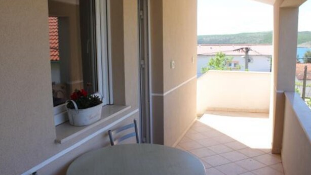 A balcony with a round table and chairs, a window with a plant, and a view of buildings and hills.