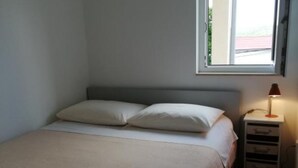A bedroom with a bed, two pillows, a nightstand with a lamp, and a window with a view of greenery outside.