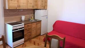 A compact kitchen with wooden cabinets, a white stove, a sink, and a refrigerator. A red sofa and a wooden chair are in the adjacent living area.