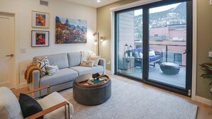 A modern living room with a grey sofa, a round ottoman, and a glass door leading to a balcony with outdoor furniture. - Big Hill Penthouse Chalet (Revelstoke)