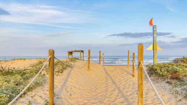 Een zandpad langs het strand, met houten palen en een touwbalk als afscheiding naar de zee.