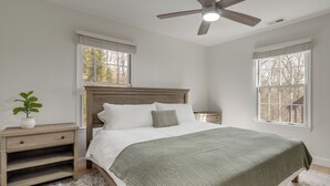 Un dormitorio con una cama, una mesita de noche, un ventilador de techo y dos ventanas.