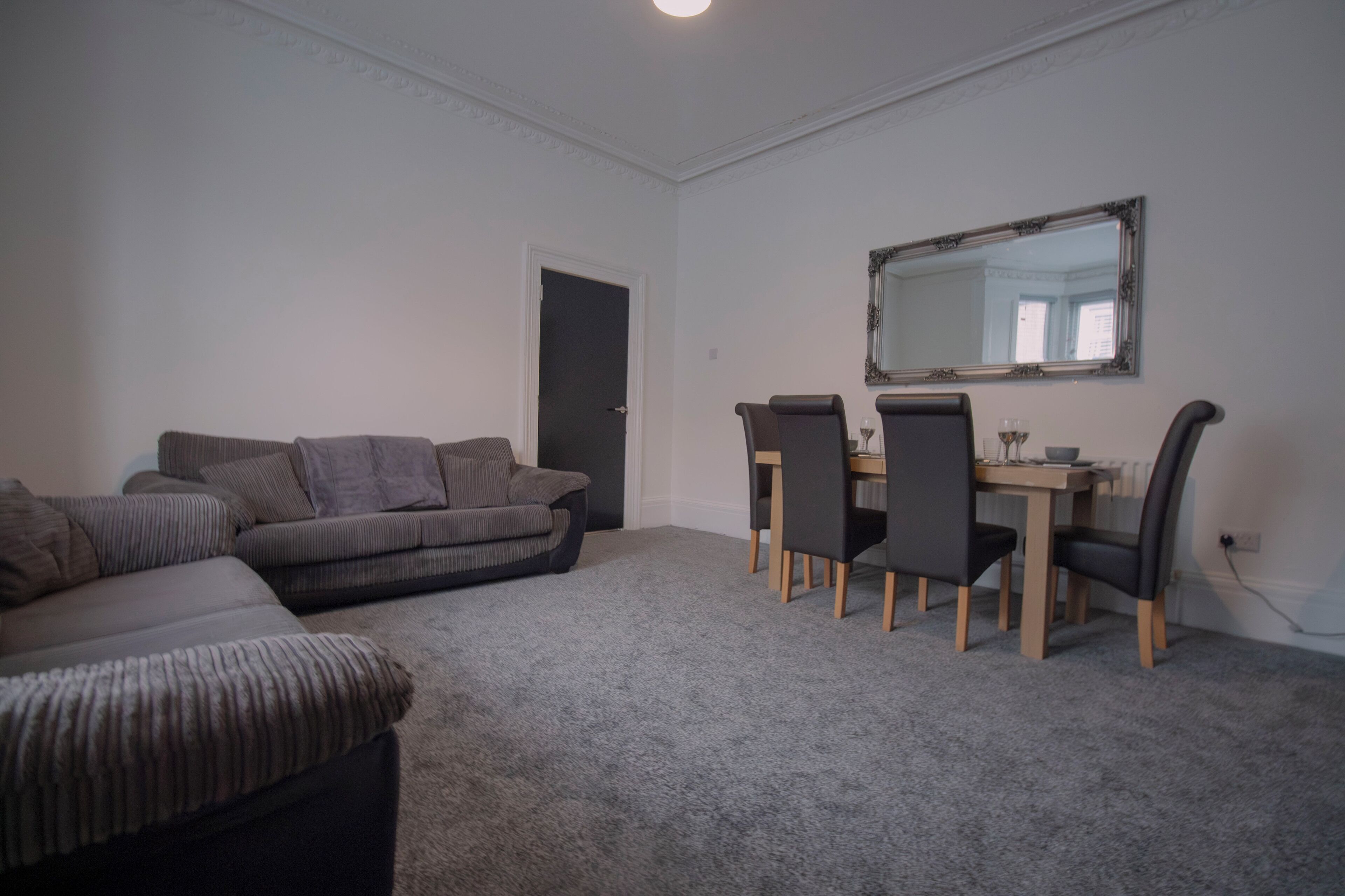 A living room with a grey sofa, a dining table with four chairs, and a mirror on the wall.