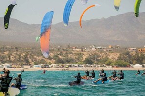 A beach with multiple kitesurfers in the water, a mountainous backdrop, and beachside buildings. - Vista Cortes 2BR 2BA Condo - Walk to the Malecon! (La Paz)