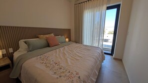 A bedroom with a bed, a nightstand, a window with curtains, and a view of the outdoors. - Vista Cortes 2BR 2BA Condo - Walk to the Malecon! (La Paz)