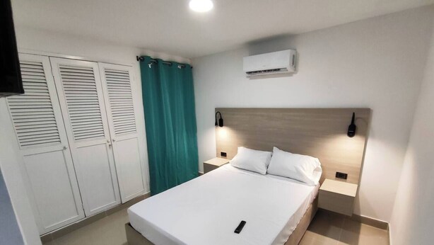 A neatly arranged bedroom with a bed, a wardrobe, a curtain, an air conditioning unit, and bedside lamps. - Edificio Los Manglares  (Cartagena)