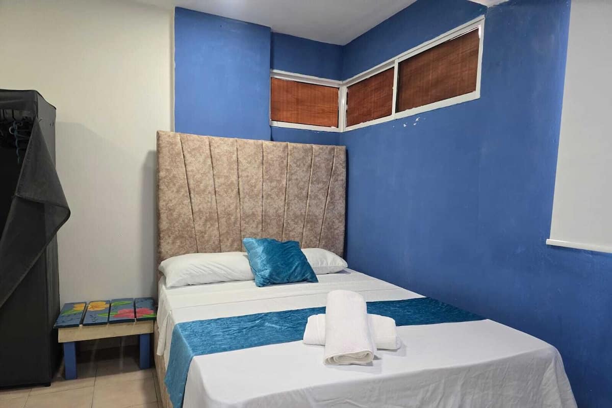 Un dormitorio con una cama, pared azul, una ventana y una mesita con un tapete colorido.