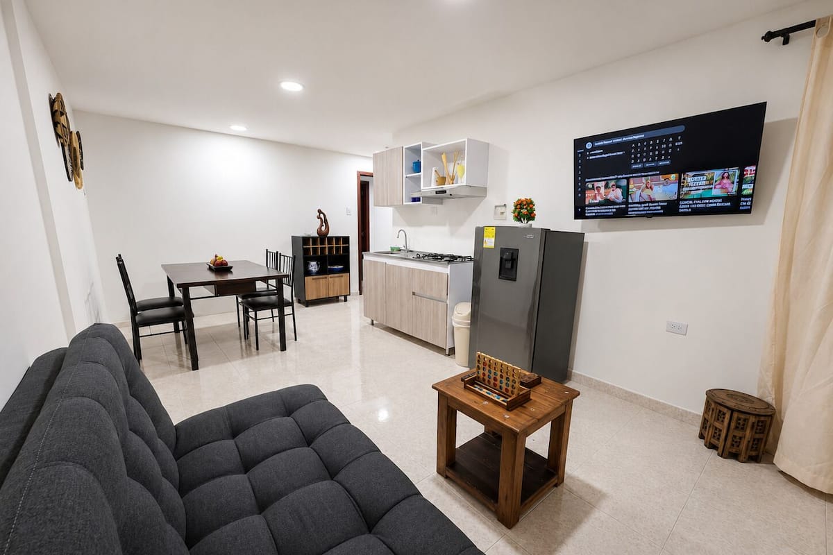 Una sala de estar moderna con un sofá, una mesa de centro de madera, una zona de cocina con armarios y un refrigerador, y un televisor montado en la pared.