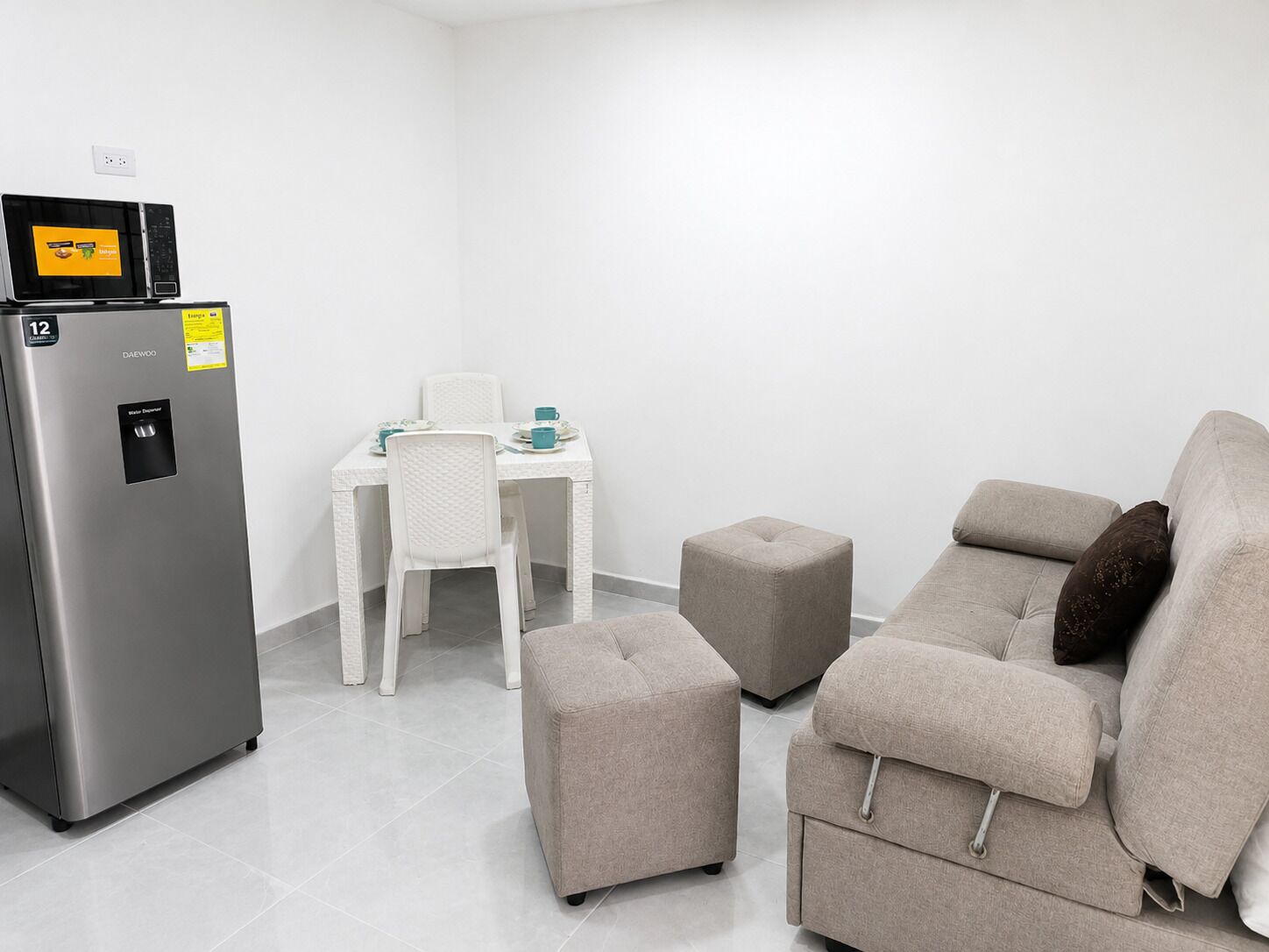 A compact living space with a kitchenette, a white dining table with chairs, a beige sofa, and a small ottoman.