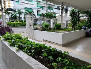 Un bâtiment moderne doté d’une entrée sécurisée par un portail, d’un bac en béton contenant des plantes vertes et d’une zone dallée.