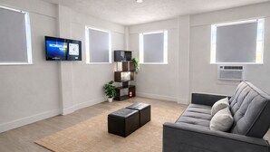 A modern living room with a grey sofa, a black ottoman, a TV mounted on the wall, and a plant on a shelf. - Charming Little Nook Near Universal Parks & Outlets. (Orlando)