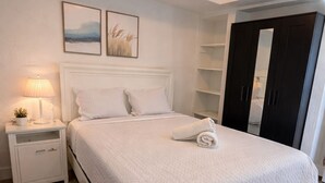 A neatly made bed with a white quilt, a bedside lamp, and a small plant on a white nightstand. There are two framed pictures on the wall above the bed. - Charming Little Nook Near Universal Parks & Outlets. (Orlando)
