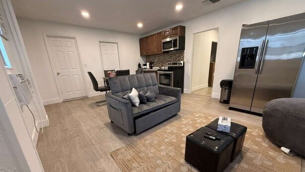 A modern living room with a grey sofa, a black ottoman, a kitchen area with a refrigerator, and a microwave. - Charming Little Nook Near Universal Parks & Outlets. (Orlando)