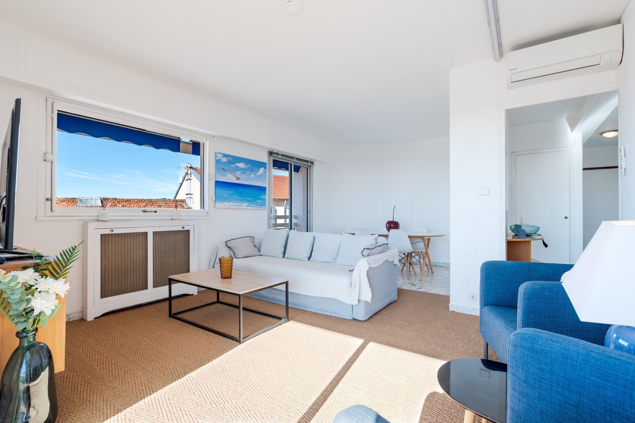 Ein modernes Wohnzimmer mit einer Couch, einem Couchtisch und einem blauen Sessel. Ein großes Fenster bietet Blick auf ein Gebäude, und an der Wand hängt ein Bild mit einer Strandansicht.