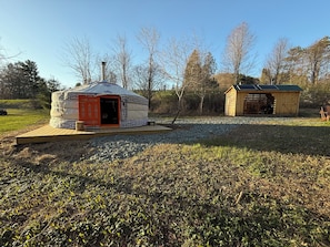 Eine Jurte mit offener Tür, ein Holzlagerhaus und ein Kiesweg.