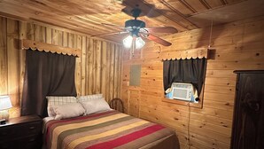 Camera da letto in una baita di legno, con un letto, un comodino, una ventola a soffitto e una finestra con tende.