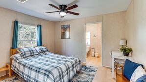 Una camera da letto con un letto, una ventola a soffitto, una finestra con tende, un quadro, una porta del bagno, un tavolino con una lampada e una sedia blu.