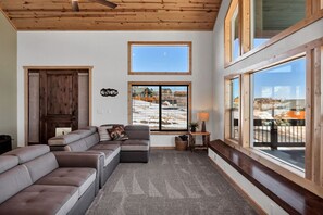 Ein geräumiges Wohnzimmer mit einer großen Eckcouch, Holzboden und großen Fenstern mit Blick auf eine verschneite Landschaft.