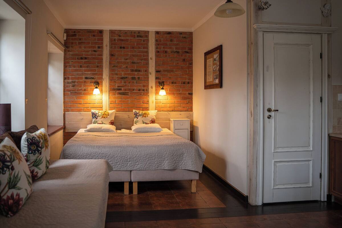 Un dormitorio con pared de ladrillo, cama con cubrecama blanco, mesita de noche, lámpara y una puerta.