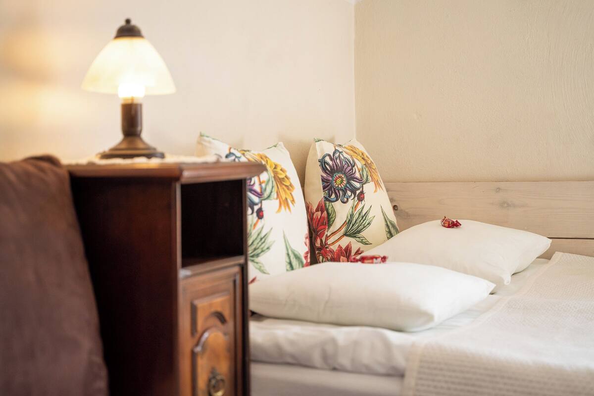 Una cama bien tendida con sábanas blancas, una mesita de noche de madera con una lámpara y almohadas estampadas con motivos florales.