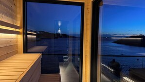 Una cabaña moderna con interior de madera, una puerta de cristal que da a un balcón con vistas al agua y un faro distante.
