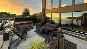 Una terraza moderna al aire libre con mobiliario, vista al agua y un edificio grande con ventanas reflectantes.