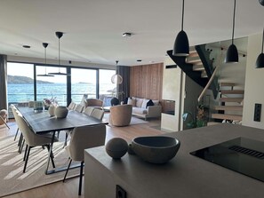 A modern interior with a dining area, seating, a staircase, and a view of the sea.