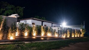 A building with a white facade and a tiled roof, surrounded by a well-lit garden with trees and a pathway leading to the entrance. - QUINTAL DE BÚZIOS
Casa com piscina, churrasqueira, 3 suítes completas com ar (Armação dos Búzios)