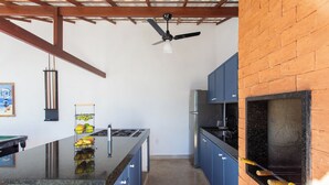 A kitchen with a central island, blue cabinets, a black countertop, a ceiling fan, and a brick wall. - QUINTAL DE BÚZIOS
Casa com piscina, churrasqueira, 3 suítes completas com ar (Armação dos Búzios)