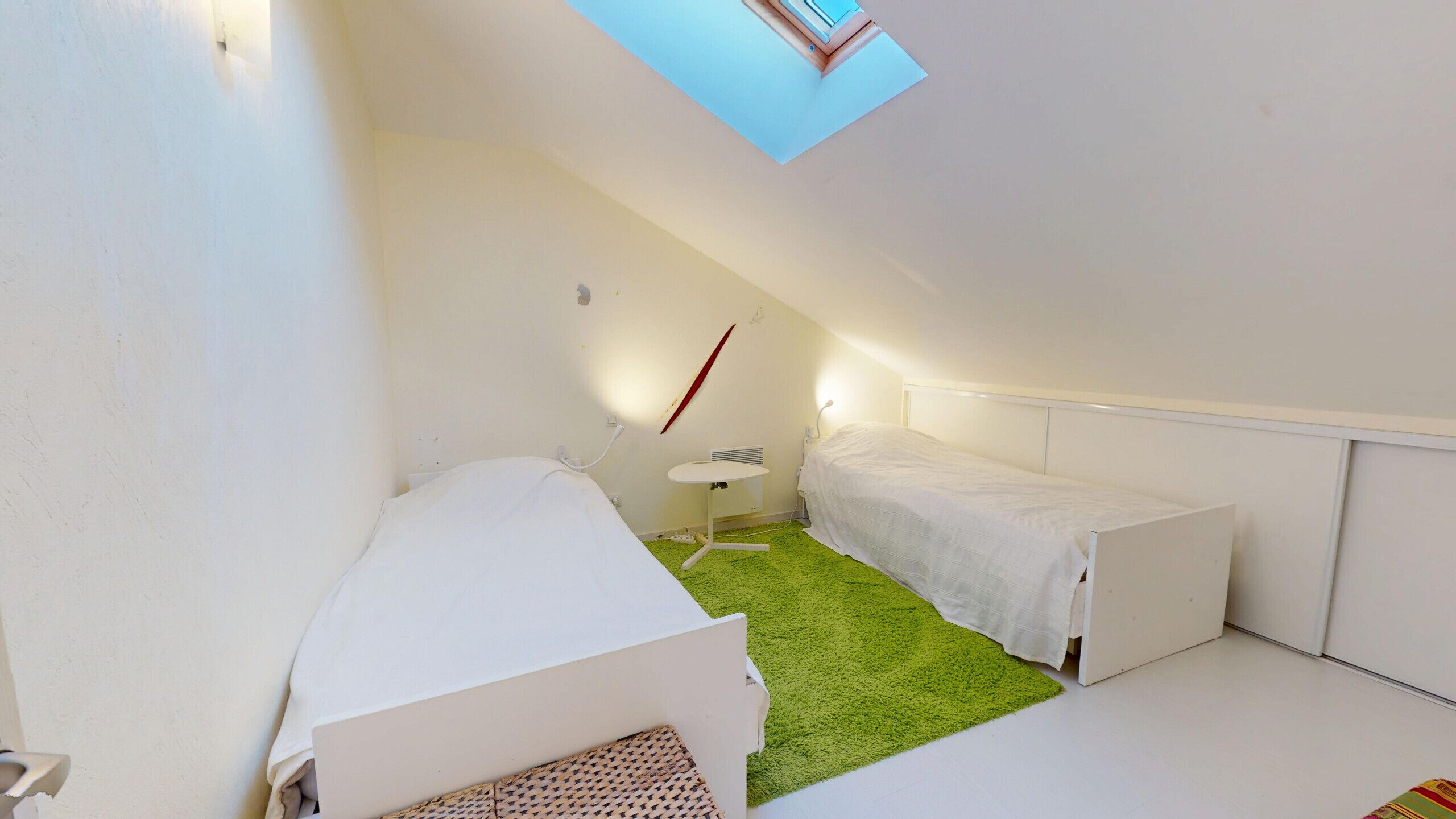 A small, white, attic-style bedroom with a green carpet, a single bed, a small table, and a slanted ceiling with a skylight.