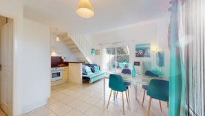 A spacious living area with a dining table, chairs, and a sofa. A kitchen is visible in the background with a staircase leading to an upper level. - Escale Bleu - House with Heated Pool (Notre-Dame-de-Monts)
