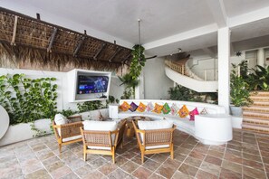 Een zonnig patio met een witte bank, rieten stoelen, een aan de muur bevestigde televisie en een trap naar een hoger niveau.