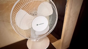 Un ventilador Ambiance blanco sobre una pequeña mesa en una habitación con una puerta de madera y una pared color beige.