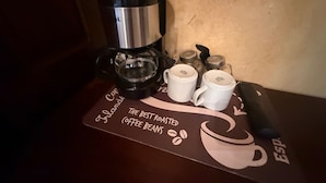 Una cafetera, dos tazas blancas y un soporte para cápsulas de café sobre una mesa con un mantel individual de temática cafetera.