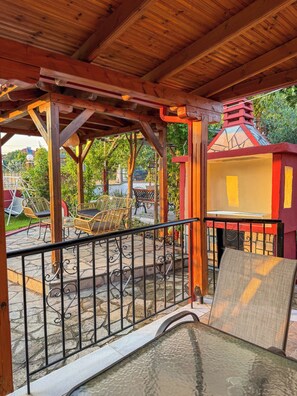 Un espacio al aire libre techado con una pérgola de madera, una pequeña estructura amarilla para juegos y una mesa de vidrio con una silla.