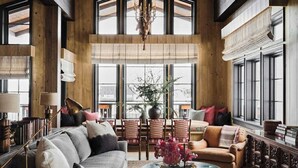 A spacious living room with a large sofa, a coffee table, and a fireplace. There are multiple windows providing natural light and a view of the outdoors.