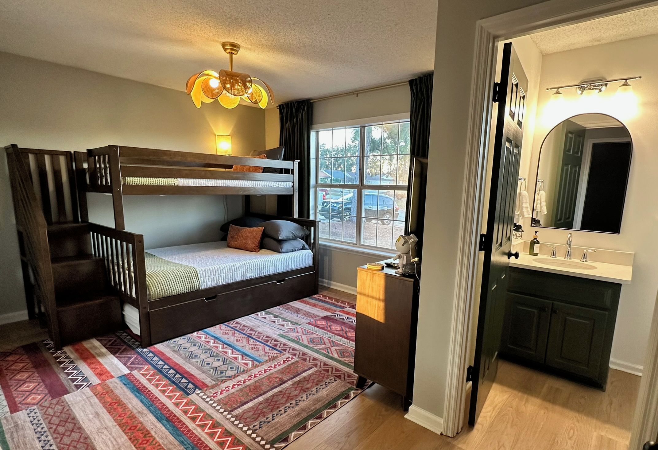 Une chambre avec un lit superposé, un bureau, un miroir et une fenêtre donnant sur des voitures garées.