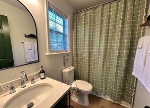 Un baño con lavamanos blanco, inodoro, ventana con persianas y cortina de ducha verde.