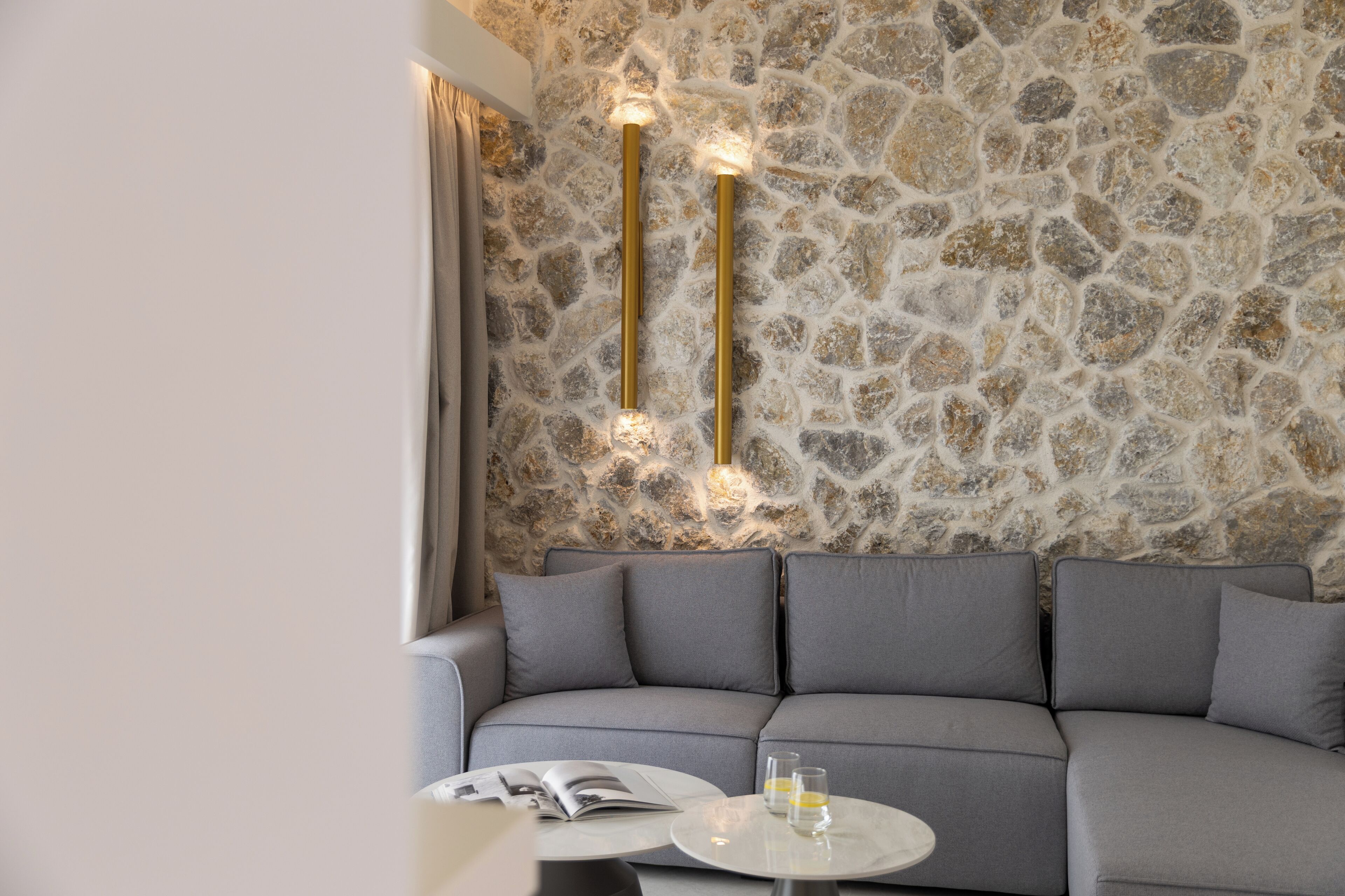 Un intérieur moderne avec un mur en pierre, un canapé gris et une petite table ronde sur laquelle sont posés des verres.