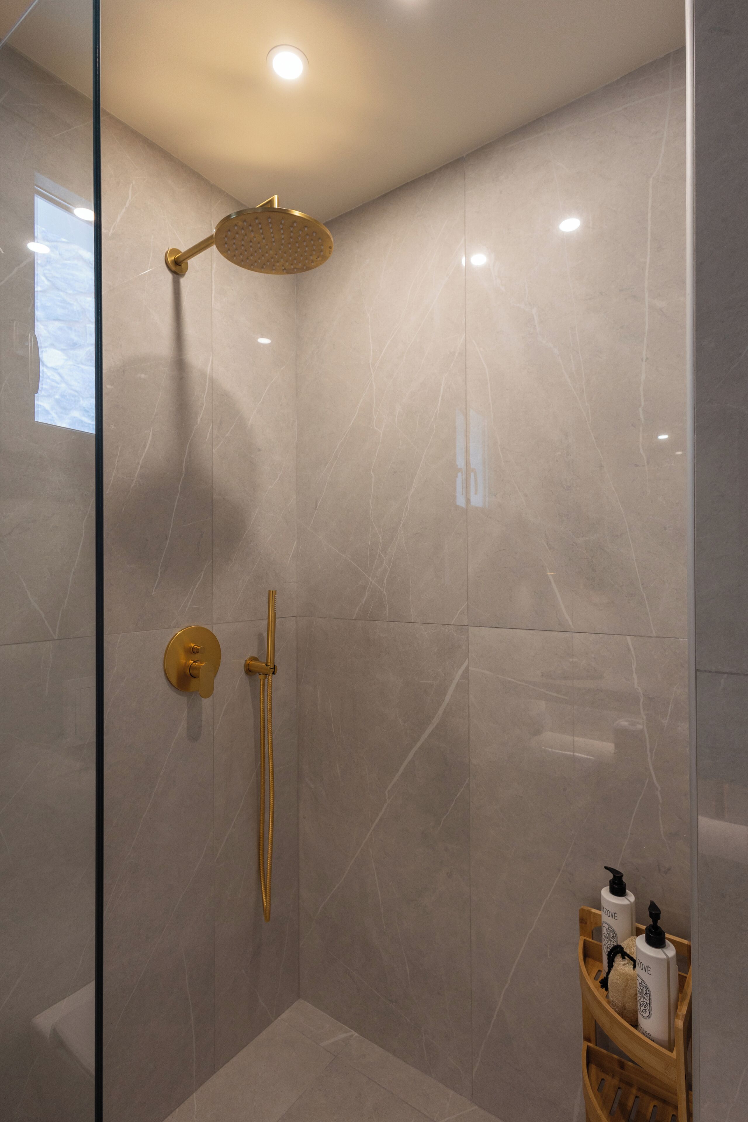 Una ducha moderna con cabezal de ducha dorado, un cabezal de ducha de mano y un estante de madera con artículos de aseo.