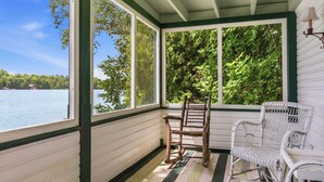 A covered porch with a view of a lake, a wooden chair, and a wicker chair. - Villa von Baumbach - Trade Screens for Stories (Elkhart Lake)