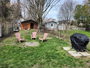 A backyard with three Adirondack chairs, a grill, and a house with a shed.