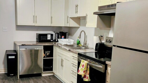 A compact kitchen with white cabinets, a stainless steel dishwasher, a microwave, a stove, and a refrigerator. - Cozy Brockville Getaway | Walk to River, Dining & Downtown (Brockville)