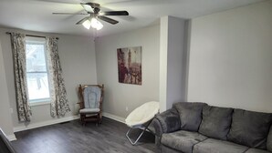 A living room with a grey sofa, a wooden coffee table, a white chair, a wooden chair, a ceiling fan, and a painting on the wall. - Cozy Brockville Getaway | Walk to River, Dining & Downtown (Brockville)