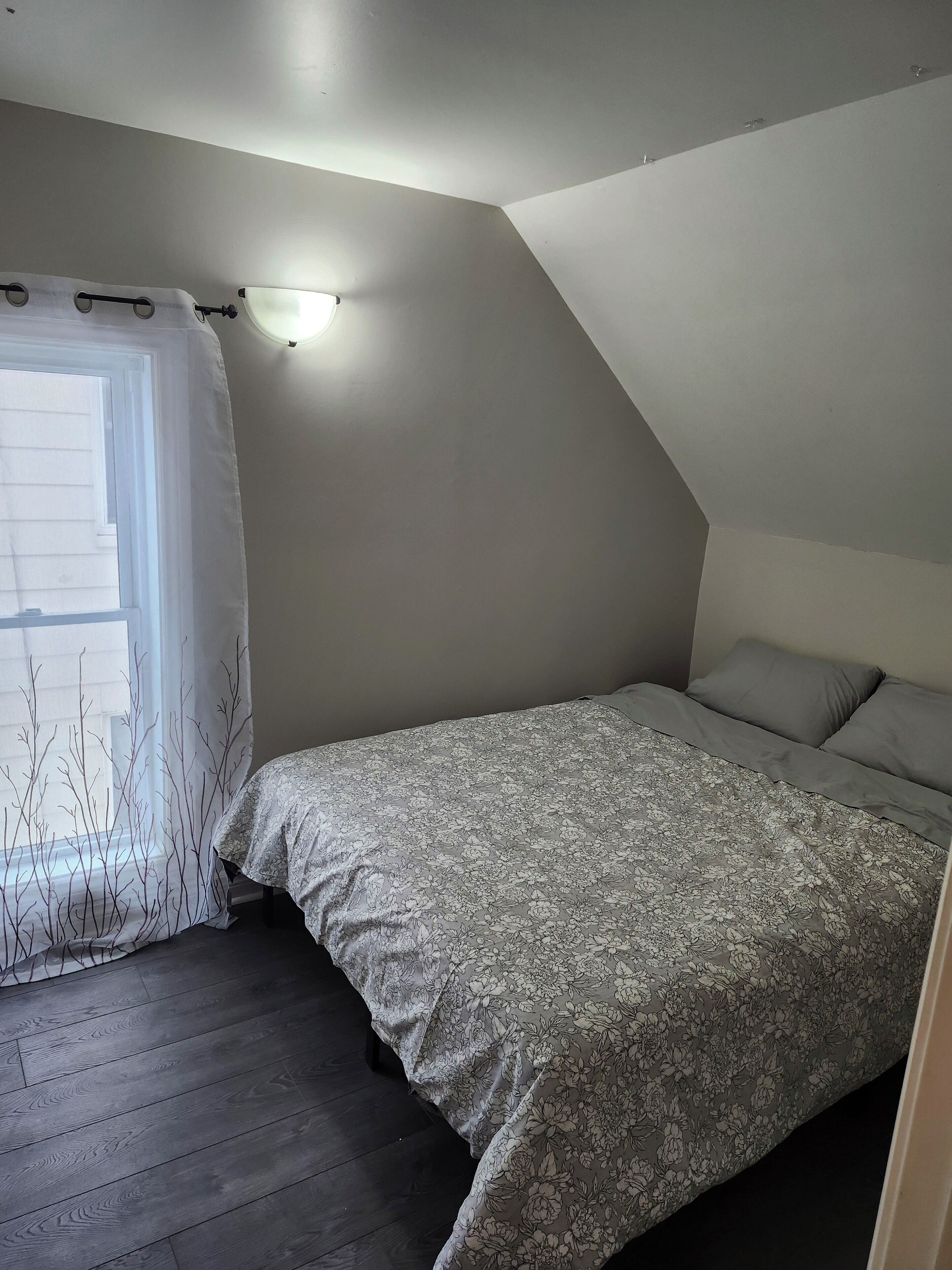 Une chambre avec un plafond en pente, un lit recouvert d’une couvre-lit à motifs floraux, une fenêtre ornée d’un rideau léger et un luminaire fixé au mur.